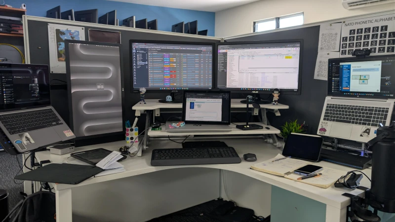 One keyboard and mouse moves across a Windows laptop, a Windows tablet, and a Mac, with Synergy keeping them all connected. The Synergy config screen is open front and center, mid-setup. A row of spare monitors lines the wall behind, hinting at the broader IT environment this desk sits in.