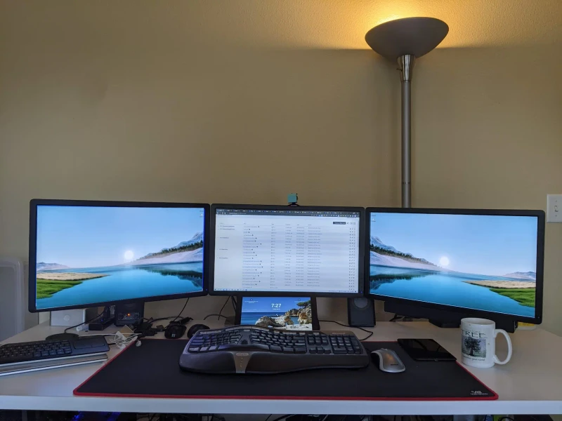 Three machines share this desk: a main PC, a work laptop across two monitors, and a Surface propped up front for video streaming. One keyboard and mouse covers all of them via Synergy, so switching between work tasks and background media takes just a nudge of the cursor.