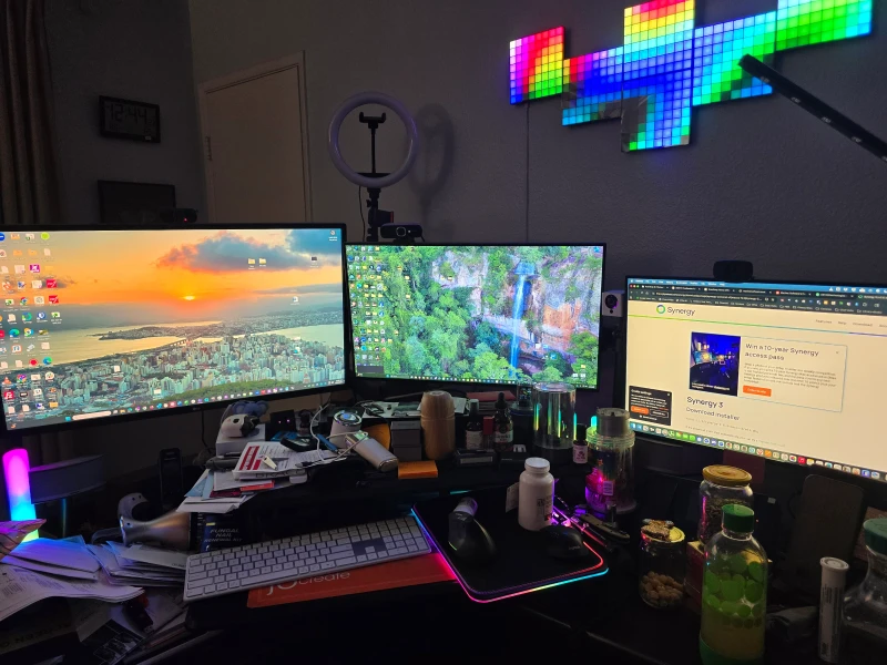 Three machines share this desk: an HP ProDesk, an HP Envy, and a Mac Mini M2, each on its own display. Synergy keeps one keyboard and mouse moving between all three, which is the only practical way to run a Windows, Windows, and macOS setup without a tangle of peripherals.