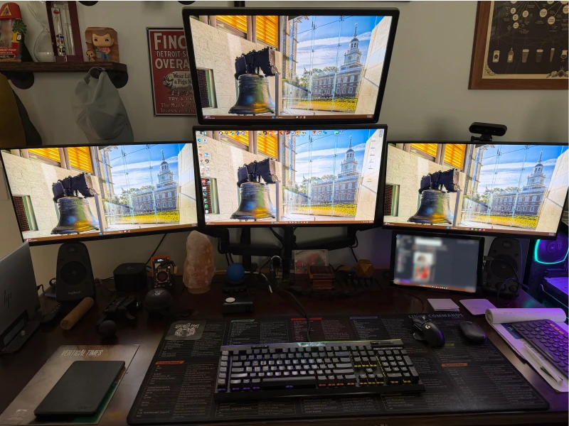 Four machines on one desk, one keyboard across all of them. The main build is a Ryzen 7 9700X with a Radeon 7900 XTX, paired with two Surface devices and an HP EliteBook, each reachable without touching a second input. A Linux commands cheat sheet sits right on the desk mat for quick reference.