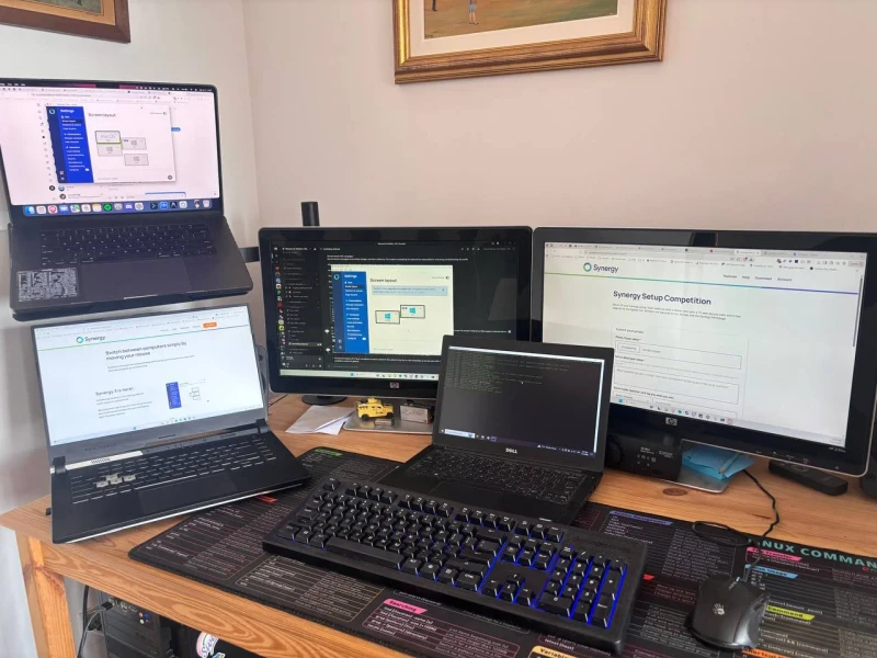 One main PC anchored to a mix of laptops, from a MacBook Pro and ASUS ROG Strix down to a small Dell and MSI, all sharing a single keyboard and mouse through Synergy. The desk mat printed with Linux commands is a practical touch for a setup that clearly spans more than one OS.