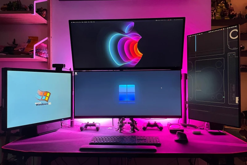 Windows 11 handles the main workload across two displays, a personal Mac sits above for everyday use, and a work Mac lives on the right side. One keyboard and mouse moves across all three operating systems through Synergy, keeping everything reachable without swapping peripherals.