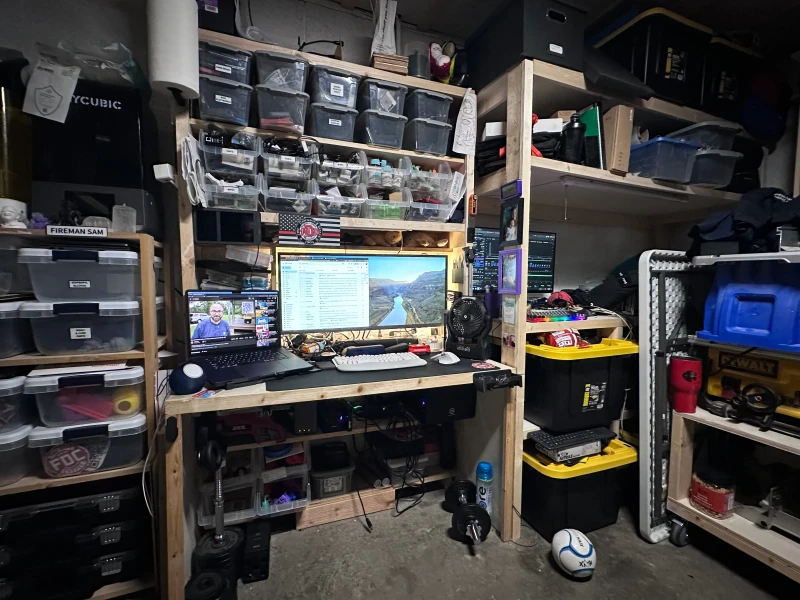 MacOS 26 on the MacBook Pro alongside Debian on the second machine, with a single Bluetooth keyboard and mouse shared between them through Synergy. The whole thing lives in a busy workshop, surrounded by labeled bins, power tools, and a 3D printer.