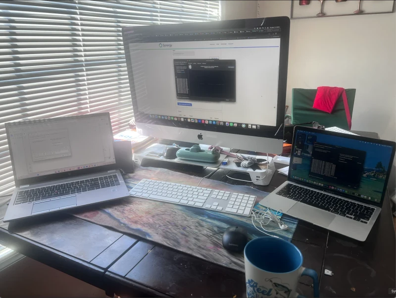 Three machines, three operating systems: a work HP ZBook on Windows, a personal iMac, and an M1 MacBook Air, all sharing one keyboard and mouse. One cursor slides between them, so there's no more stretching to reach a laptop touchpad or adding extra peripherals to an already busy desk.
