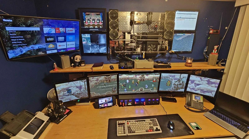 The main PC mounted to the wall drives five displays, while a Zima board, a Lenovo Legion Go running Bazzite, and a Framework laptop on Xubuntu all share the same keyboard and mouse through Synergy. Four very different operating environments, one input set sliding between them.