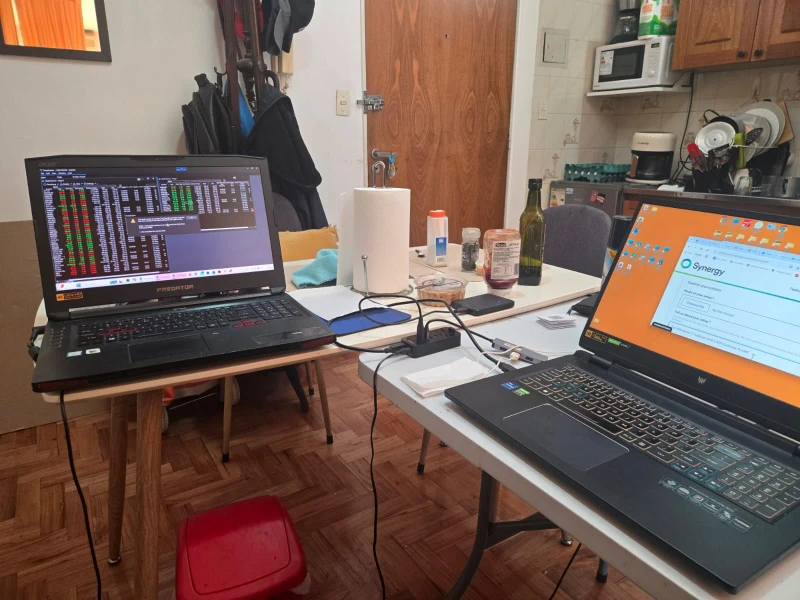 Two laptops and a WiFi gateway router, all sharing one mouse and keyboard through Synergy. One machine runs what looks like a busy terminal dashboard while the other handles browsing, both reachable without switching peripherals.