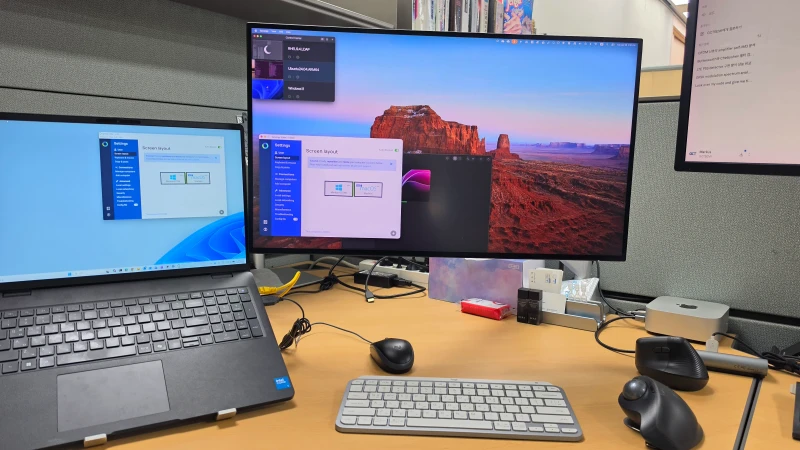 Synergy connects the Windows 11 laptop and M4 Mac Mini here, letting one keyboard and mouse move between them across platforms. The Synergy screen layout is open on both machines, showing the setup mid-configuration at the cubicle desk.
