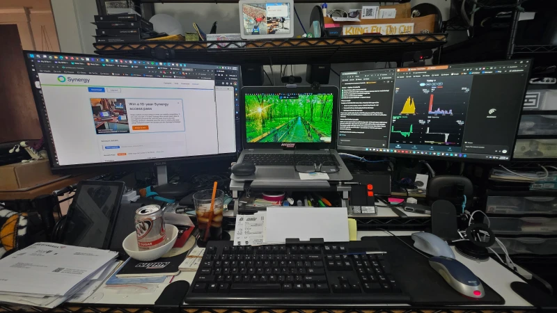 One keyboard and mouse covers three machines here: two Windows 11 desktops, one Intel i5 and one AMD Ryzen 5, plus a laptop, all linked through Synergy. The well-used desk has a lived-in energy, papers and notes filling every gap between the machines.