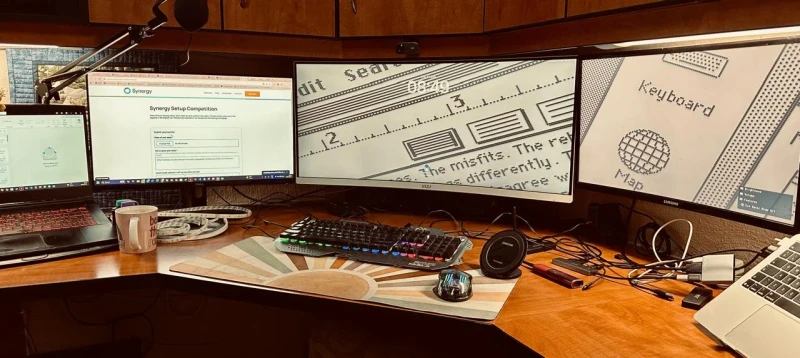 One keyboard and mouse moving between a Windows laptop and a MacBook Air, both in reach on the same curved desk. The screensaver running on the center and right displays shows the two machines are idle and ready whenever needed.