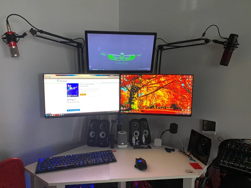 No description was provided with this entry, so there's little to go on beyond the photo itself. Three computers share this desk, with Synergy open on the left display and two condenser microphones on boom arms suggesting audio work plays a regular role here.